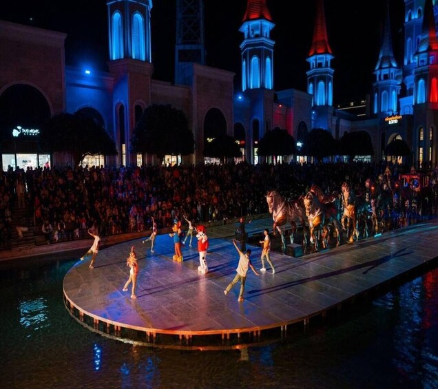 A nighttime outdoor performance with dancers on a stage, a crowd, and illuminated buildings in the background. 3