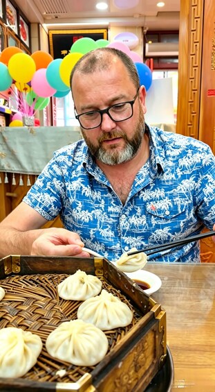 A person in a blue patterned shirt preparing food, with a basket of dumplings and a bowl of sauce on the table. 1