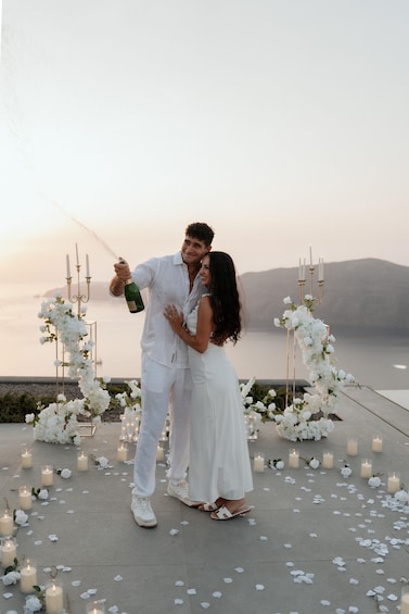 A couple embracing in a white wedding aisle with candles and flowers, overlooking a body of water. 1