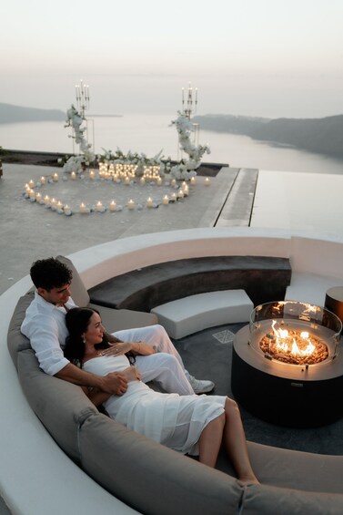 A couple relaxing on a curved white sofa by a fire pit overlooking a body of water, with a candle-lit arch and a serene landscape in the background. 4