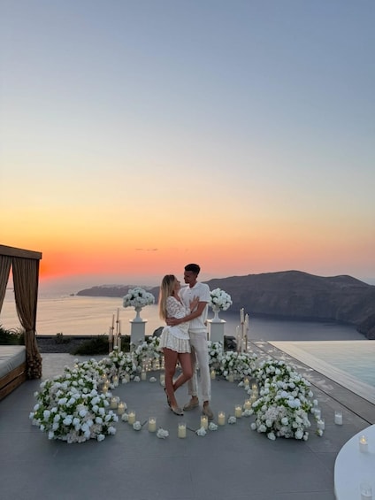 A couple embracing in a white floral arrangement on a rooftop overlooking a body of water at sunset. 1