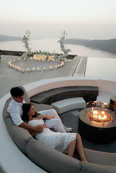 A couple relaxing on a curved white sofa by a fire pit overlooking a body of water, with a candle-lit arch and a serene landscape in the background. 3