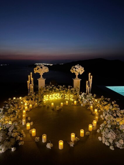 A romantic outdoor setting with a circular arrangement of candles and flowers, a backdrop of a twilight sky, and a body of water in the distance. 2