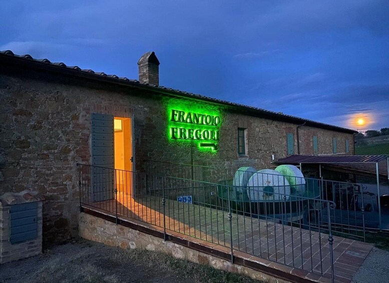 A stone building with a neon sign reading 'FRANTO FREGOI' and a fenced area with green barrels. 2