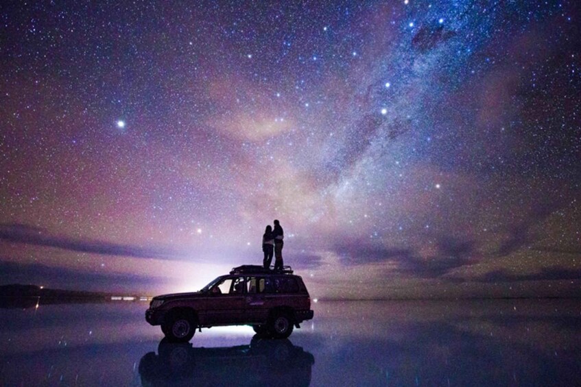 A car parked on a reflective surface with two people standing on it under a starry night sky. 3