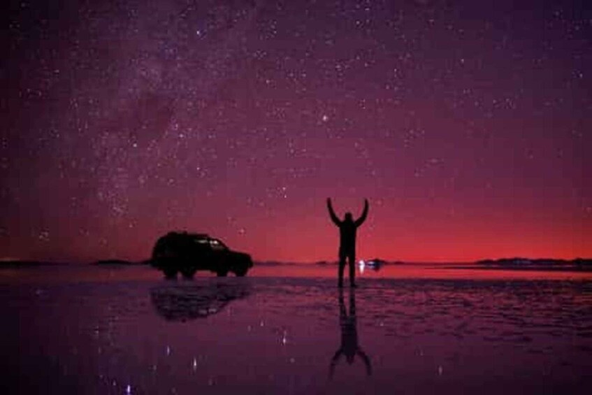 A person standing with arms raised, a car, and a starry night sky over water. 2