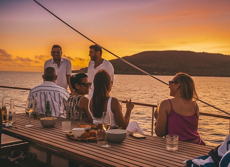 A group of people dining on a wooden boat at sunset with a view of the water and a distant mountain. 1