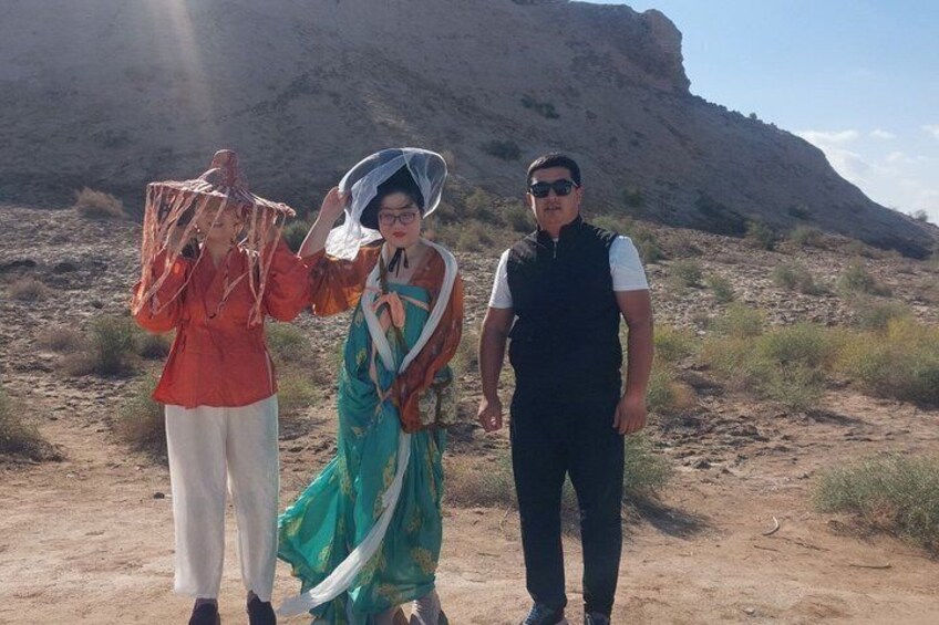 Three people in a desert landscape with sparse vegetation and a large rock formation in the background. 3