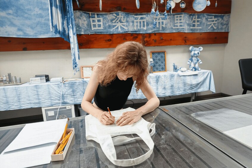 A person writing on a piece of paper at a table with a white tote bag, a box of pencils, and a blue and white patterned tablecloth. 5
