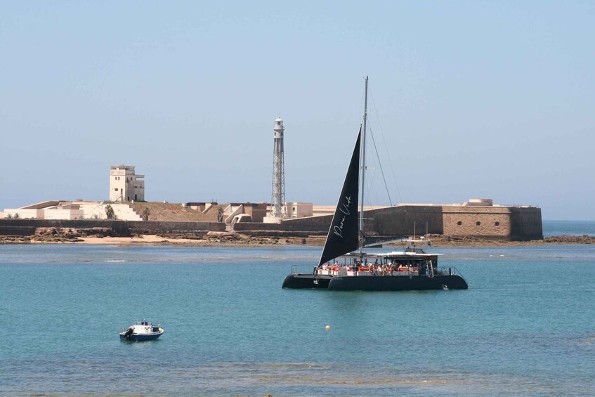 A sailboat with a black sail and a white cabin, anchored near a coastal fortification with a lighthouse. 5
