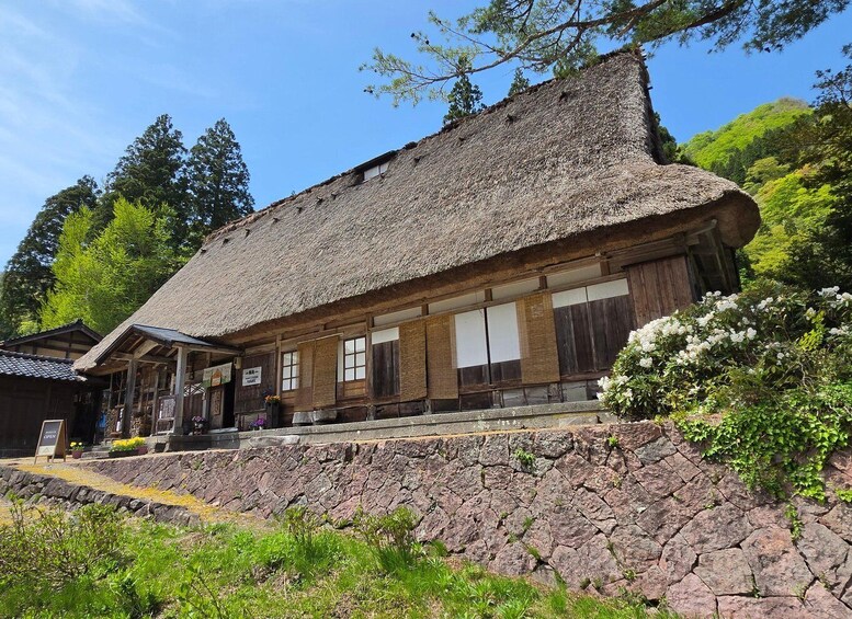 A traditional wooden building with a thatched roof, surrounded by greenery and a stone wall. 2