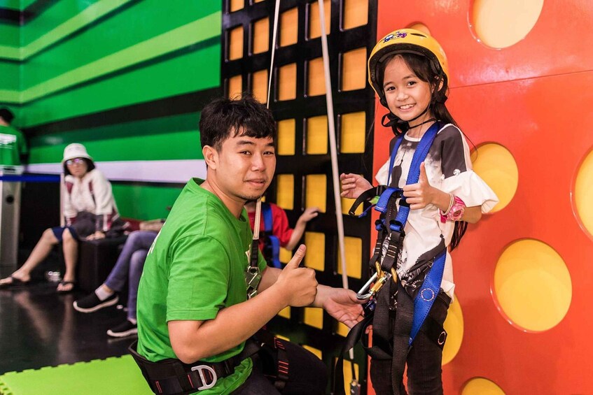 A person in a green shirt assisting another person in a white shirt with a yellow helmet, both wearing safety harnesses, in an indoor activity area with green and yellow walls. 4