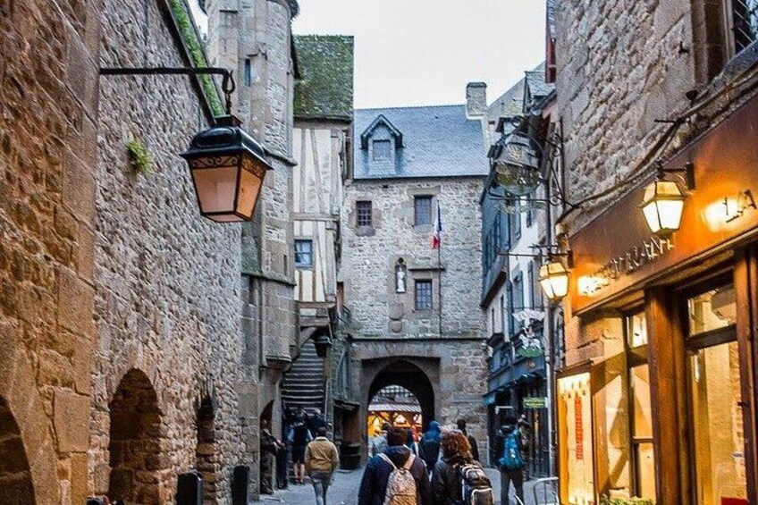 A narrow medieval street with stone buildings, a central archway, and people walking. 2