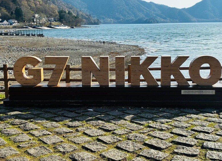 A wooden sign with 'G7 NIKKO' on a cobblestone path by a lake. 2