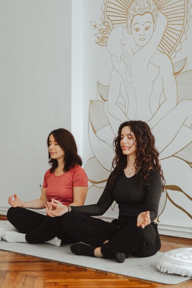 Two people sitting cross-legged on a yoga mat in a room with a large lotus flower mural on the wall. 1