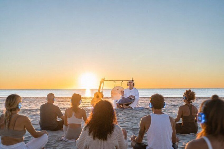 A group of people sitting on a beach at sunset, facing a person playing a guitar. 1