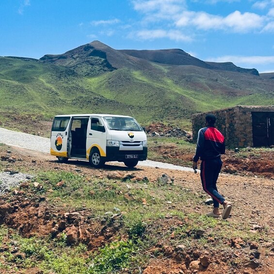 A white van with a logo on the side parked on a dirt road, a person walking towards it, and a stone building in the background. 3