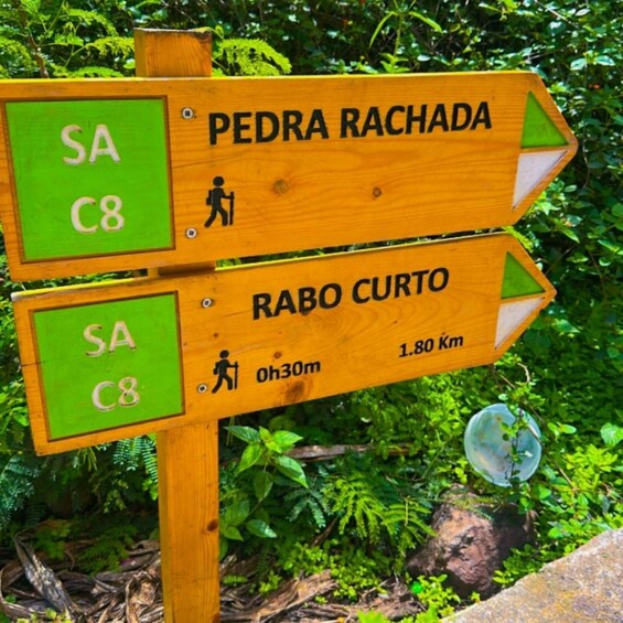 A wooden directional signpost with two arrows, one pointing to Pedra Rachada and the other to Rabo Curto, with distance markers and a walking symbol. 1