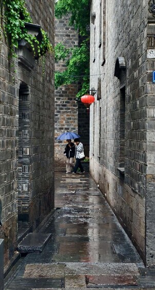 A narrow alley with wet stone walls, a red lantern, and a person with a blue umbrella. 3