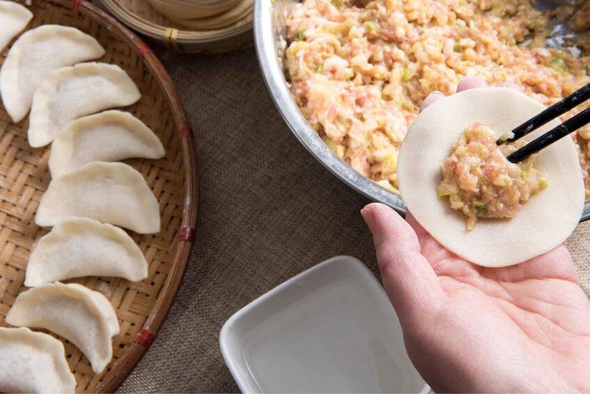 A hand holding a dumpling with chopsticks, a basket of dumplings, and a bowl of filling. 2