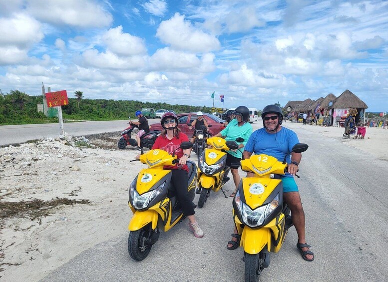 Three people on yellow motorcycles on a road with a beach and thatched-roof structures in the background. 1