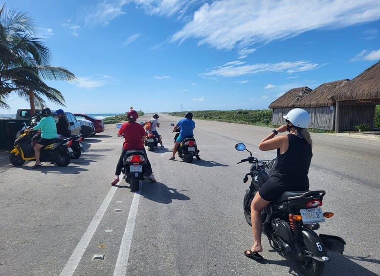 A group of motorcyclists on a road with a palm tree and a thatched-roof structure in the background. 2