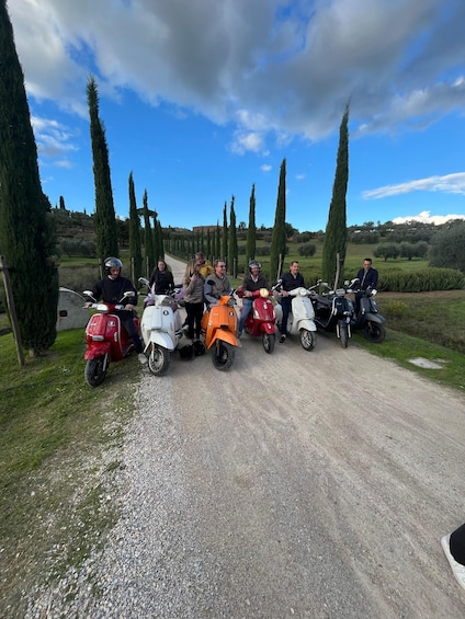 A group of people on scooters parked on a gravel road with cypress trees and a clear sky in the background. 2