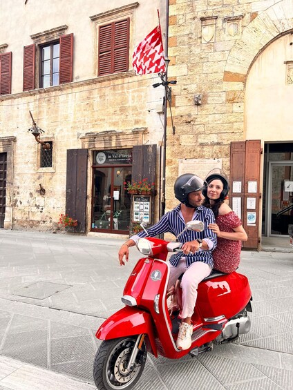 A couple on a red scooter with a flag, in front of a building with a sign that reads 'in Val d'Oc.' 5