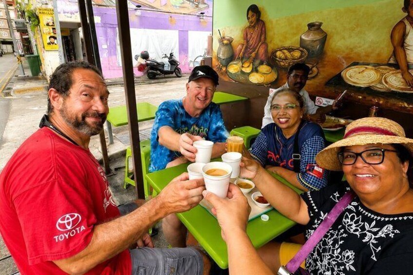 People toasting with cups at a street-side food stall with a mural in the background. 1