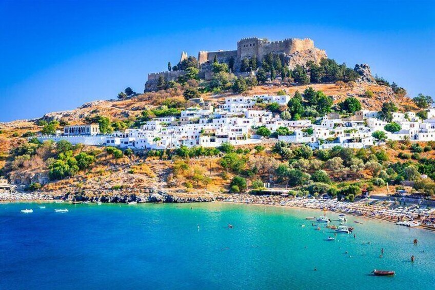 A coastal town with white buildings, a castle on a hill, and clear blue waters. 1
