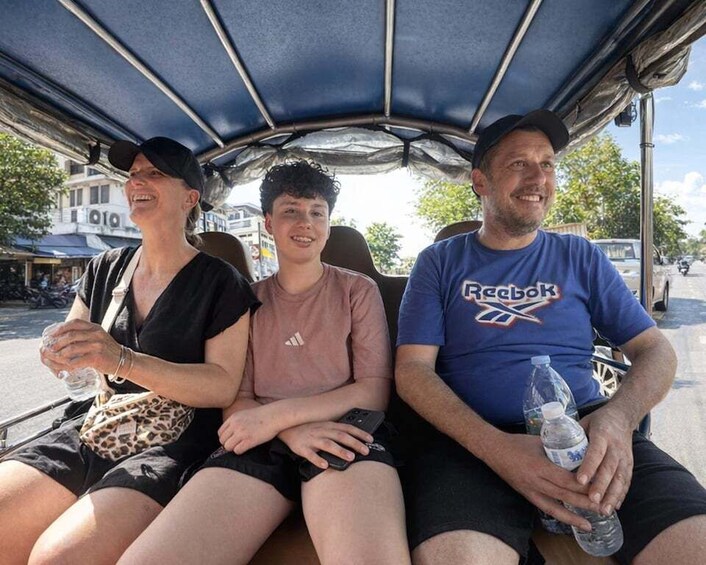 Three people seated in a covered vehicle, with a city street and buildings in the background. 3