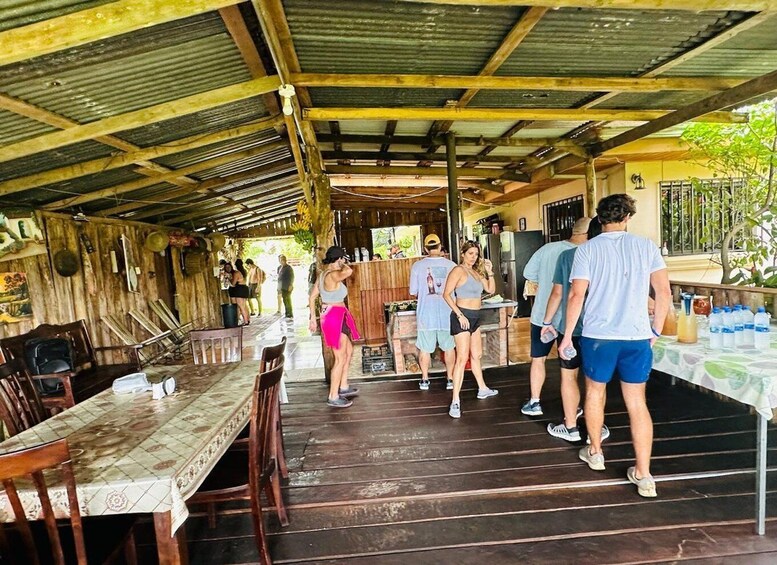 A covered outdoor area with wooden tables and chairs, a stage, and a group of people standing and conversing. 5