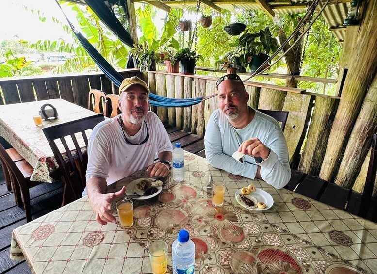 Two men dining at an outdoor table with a tropical backdrop. 4