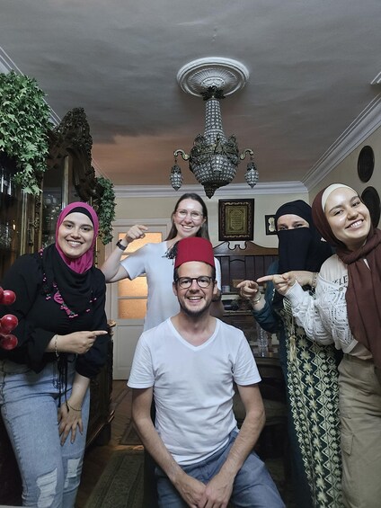A group of people in a room with a chandelier, a framed picture on the wall, and a plant. 1
