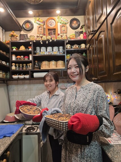 Two people holding a baked dish in a kitchen with shelves of jars and a clock on the wall. 3