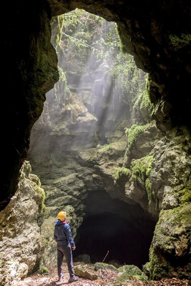 A person in a blue jacket and yellow helmet standing at the entrance of a cave with moss-covered walls and a narrow opening. 2