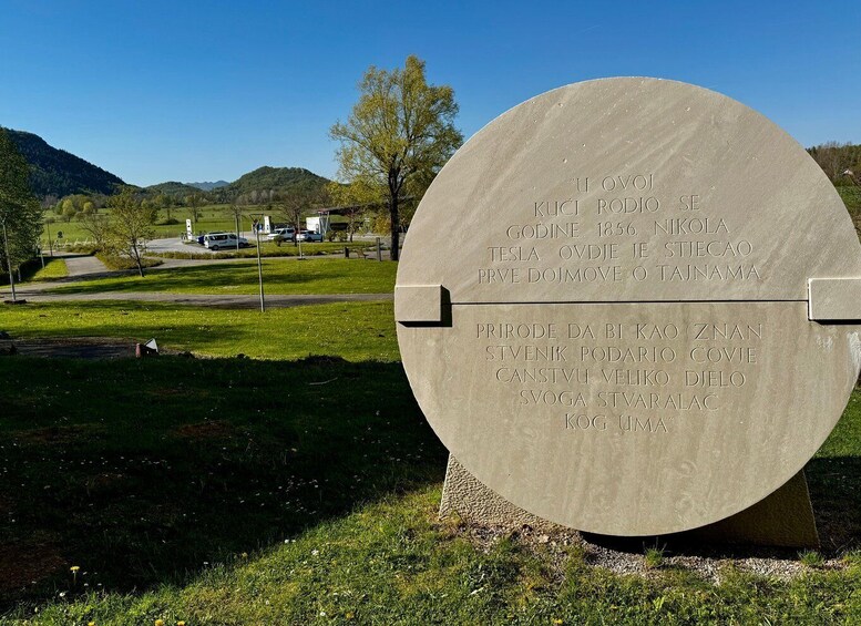 A circular stone monument with inscriptions in a foreign language, surrounded by a grassy area with trees and a mountain in the background. 3