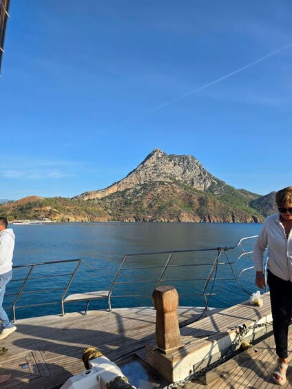 A wooden boat deck with a person walking, a mountain in the background, and calm water. 3