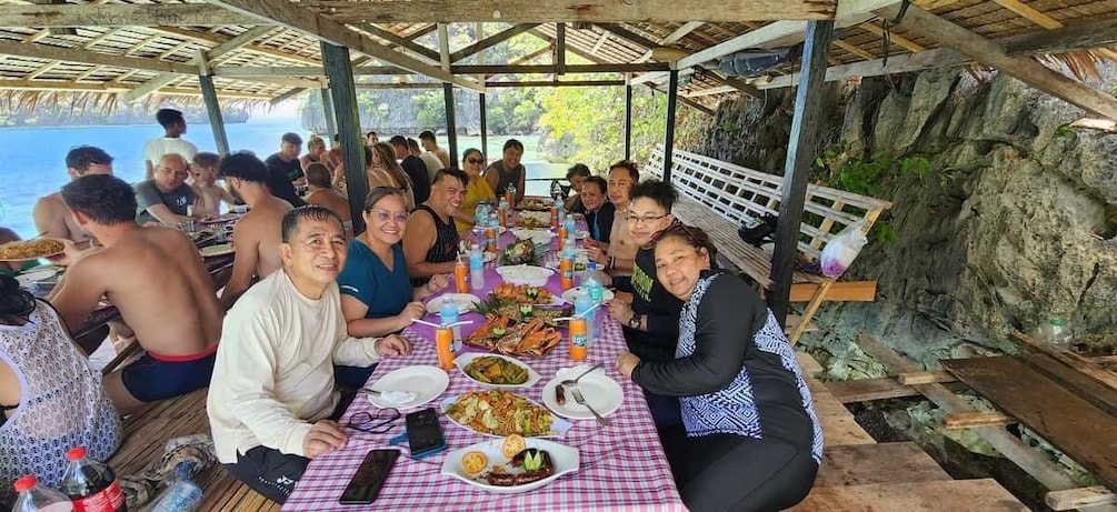 A group of people dining at an outdoor table with a checkered tablecloth, surrounded by a rocky landscape and a body of water. 5