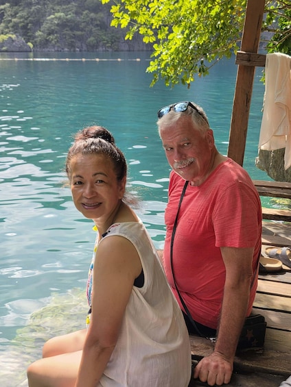 Two people sitting on a wooden dock by a calm lake with trees in the background. 2