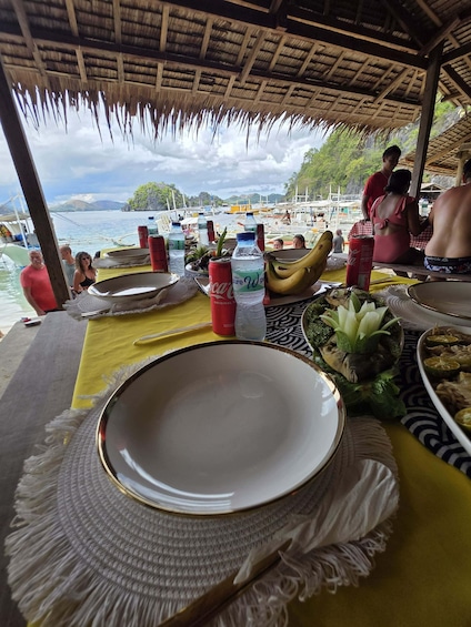 A dining table set with plates, a banana, a Coca-Cola can, and a bottle of water, overlooking a beach with boats and a thatched roof structure. 3