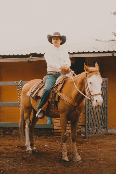 A person in a cowboy hat and boots riding a horse with a saddle, standing in front of a stable. 2