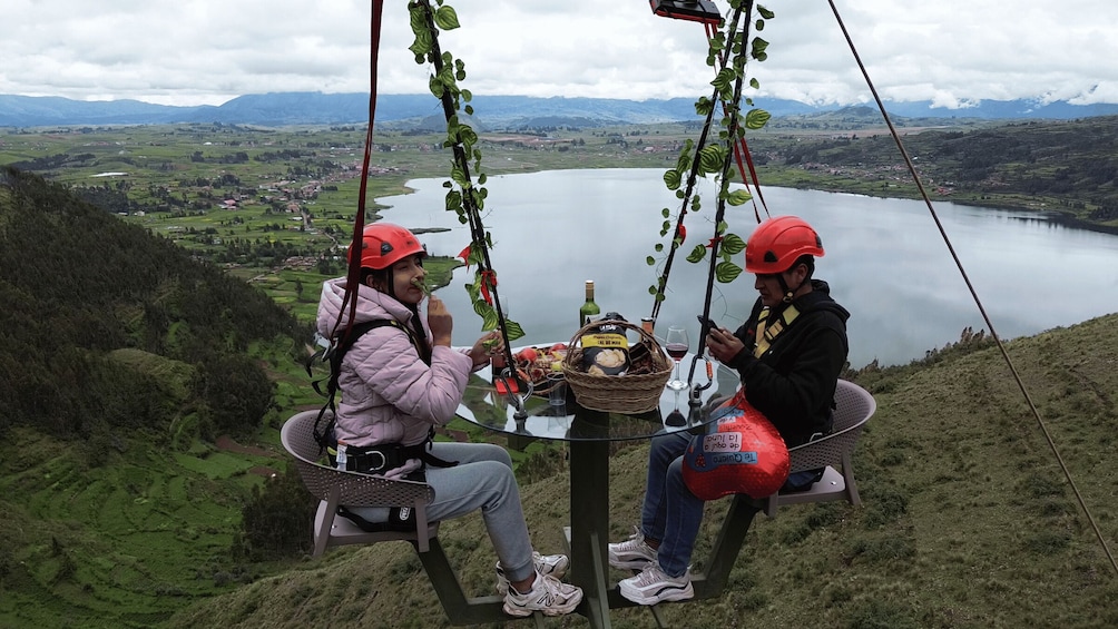 Two people on a suspended chair with a table between them, overlooking a lake and mountains. 4