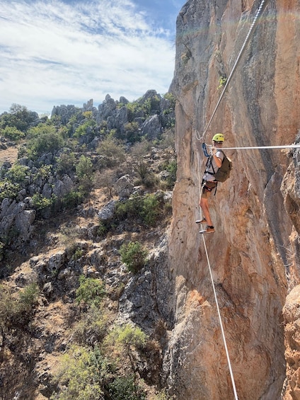 A person rock climbing a steep cliff with a harness and helmet, surrounded by rocky terrain and sparse vegetation. 1