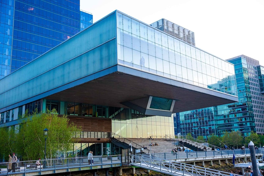 A modern building with a large overhang, glass windows, and a bridge with people walking. 1