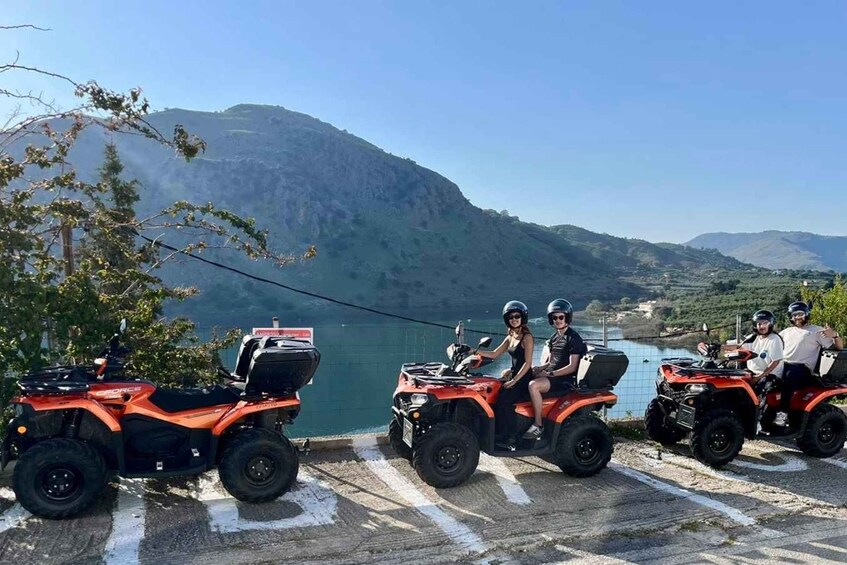Four people on ATVs parked by a lake with mountains in the background. 4