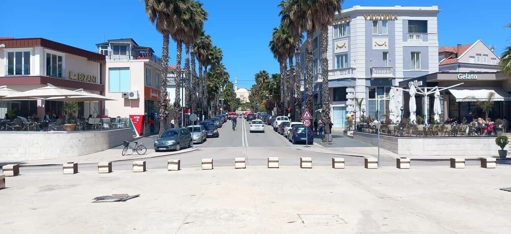 A sunny street with palm trees, parked cars, and outdoor cafes. 1