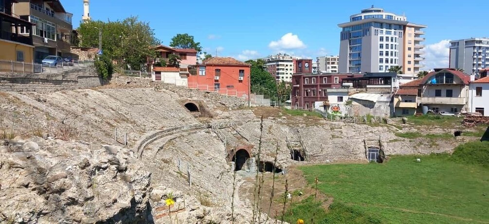 A rocky amphitheater with a clear sky, modern buildings, and a grassy area. 2