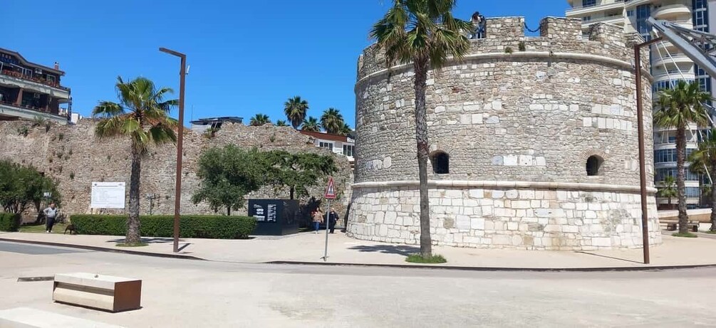 A stone tower with palm trees, a bench, and modern buildings in the background. 3