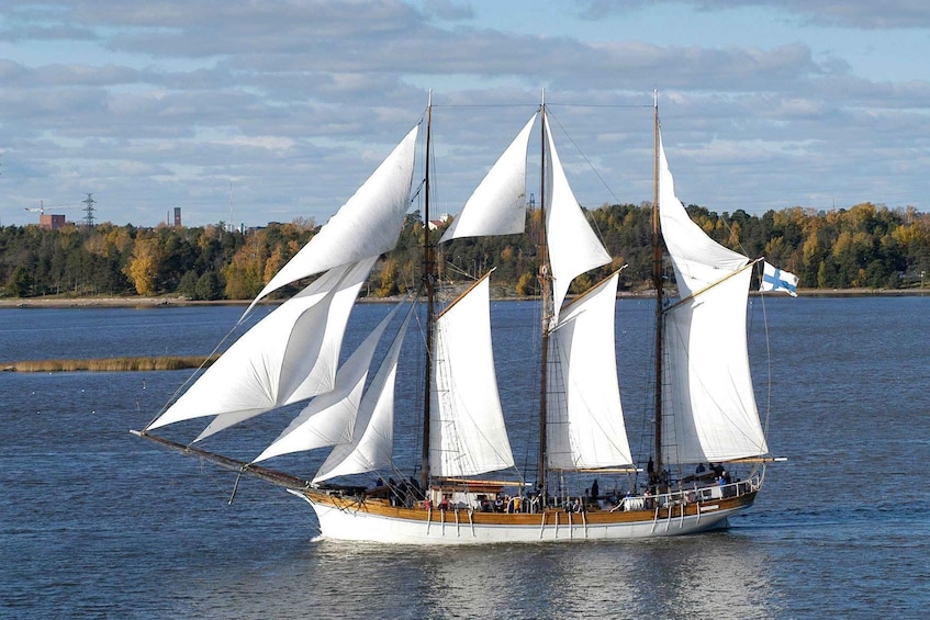 A sailboat with multiple sails on a calm body of water, surrounded by trees and a distant shoreline with buildings. 1